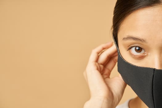 Half face shot of young woman putting on black facial mask, staying safe while posing isolated over beige background. Safety, pandemic concept