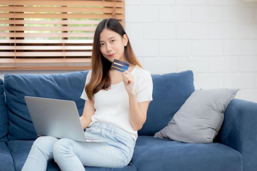 Young asian woman sit on sofa using laptop computer shopping online with credit card buying to internet, happy girl payment with e-business on couch, purchase and payment, business concept.