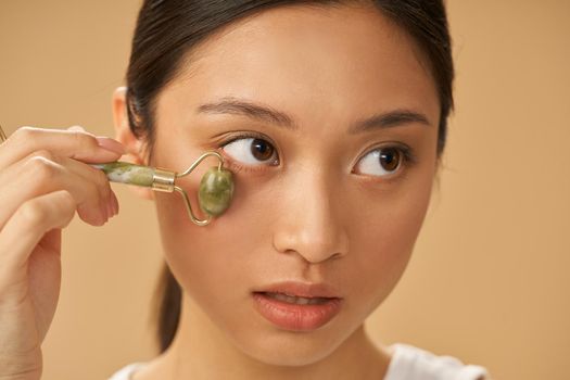 Portrait of young woman looking aside while using jade roller for massaging her face, posing isolated over beige background. Skincare concept