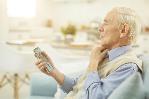 Side view of old-aged man sitting on sofa in the living room at home and using mobile phone during a consultation with a therapist