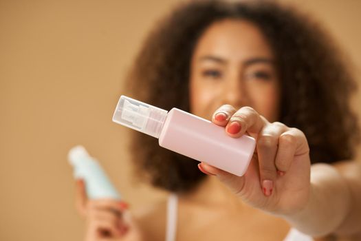 Close up of young mixed race woman holding pink bottle with beauty product while posing isolated over beige background. Focus on a bottle. Skincare concept. Horizontal shot