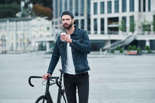 Outdoors leisure. Young stylish man walking on city street with bicycle holding cup drinking hot coffee thoughtful