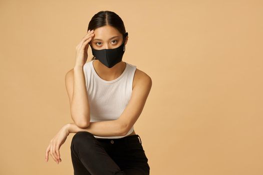 Confident young mixed race woman wearing black facial mask, staying safe, looking at camera while posing isolated over beige background. Safety, pandemic concept