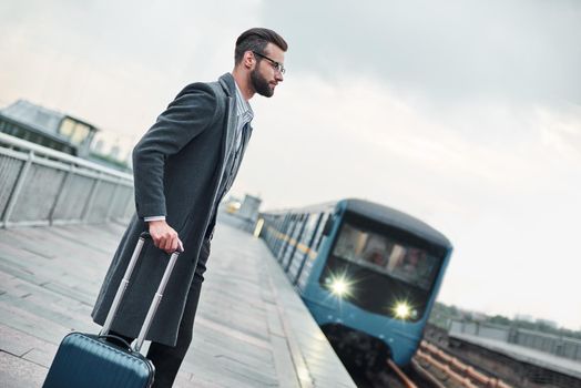 Business trip. Young businessman standing near railway with luggage waiting for train