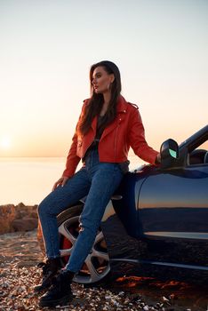 Beautiful girl near the roofless car. Autumn season, red coat and jeans, stylish sunglasses on head