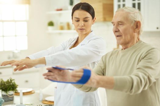 Waist up of medical professional standing near elderly man and showing exercises with fitness rubber band