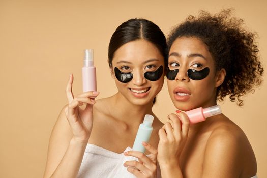 Portrait of two funny young women holding bottles with beauty product, posing with applied black hydro gel under eye patches isolated over beige background. Skincare concept