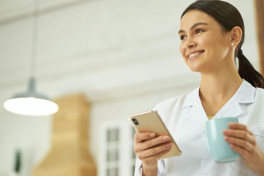 Pretty young nurse holding cup of drink and using mobile phone while standing in room