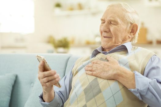 Sick Caucasian elderly man sitting on cozy sofa and using smartphone while talking online