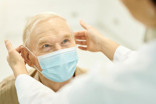 Cropped photo of medical worker in white labcoat putting a surgical mask on elderly man