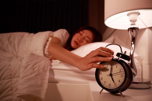 sleepy young woman in bed with eyes closed extending hand to alarm clock in the morning