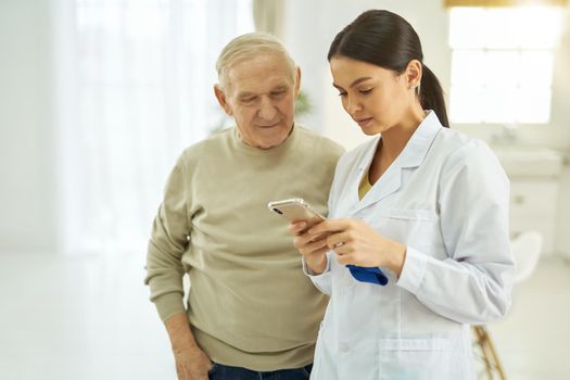 Female doctor holding the smartphone and standing near the male patient at his home