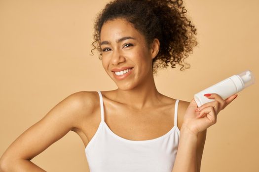 Studio portrait of cheerful young woman holding a bottle of foam facial cleanser, posing isolated over beige background. Beauty products and skin care concept