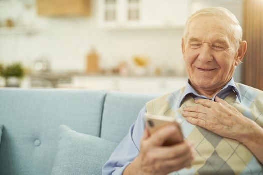 Happy Caucasian elderly man sitting on cozy sofa and using smartphone while talking online