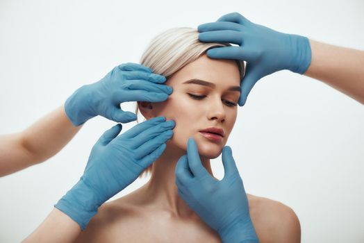 Before facial surgery. Beautiful blonde woman keeping eyes closed while doctors in blue medical gloves examining her face. Plastic surgery concept. Healthcare. Beauty concept.