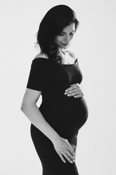 Beautiful young pregnant woman embracing belly posing in studio, black and white photo. Happy pregnancy and motherhood. High quality photo