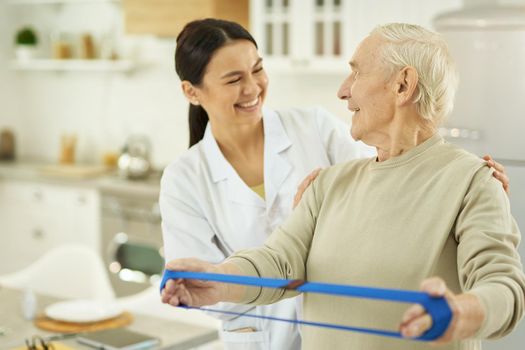 Happy senior citizen doing exercises and holding a fitness elastic band in hands while talking to female doctor