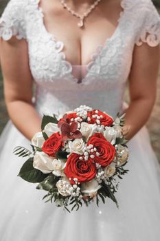 A bride in a wedding dress holds a decorative wedding bouquet of flowers of red and white roses in her hands, close-up.