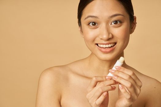 Portrait of lovely young woman smiling while holding lip balm, posing isolated over beige background. Beauty treatment concept