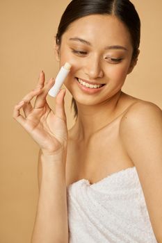 Attractive young woman wrapped in towel smiling and holding lip balm while posing isolated over beige background. Beauty treatment concept