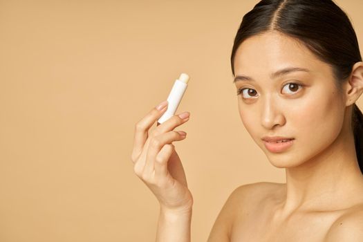Close up portrait of beautiful young woman looking at camera while holding lip balm, posing isolated over beige background. Beauty treatment concept