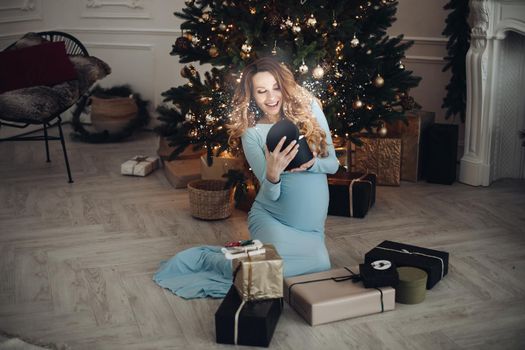 Happy pregnancy woman opening holiday light gift having positive emotion full shot. Smiling beautiful elegant pregnant girl enjoying celebrating Christmas holiday surrounded by festive decoration