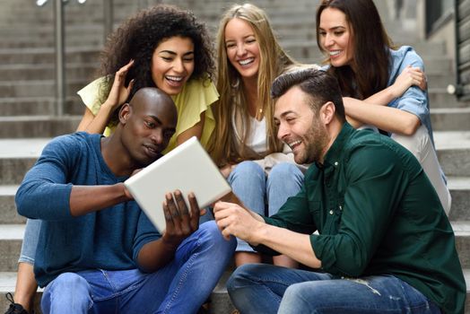 Multi-ethnic group of young people looking at a tablet computer outdoors in urban background. Group of men and woman sitting together on steps.