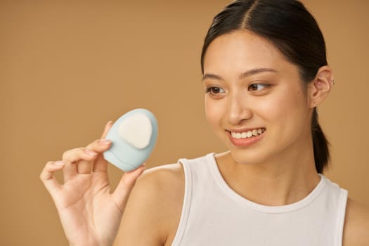 Cheerful young woman smiling and holding silicone facial cleansing brush, posing isolated over beige background. Skincare concept