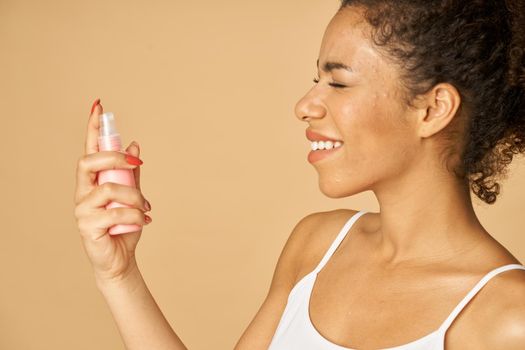 Portrait of attractive young woman applying spray water on face, posing isolated over beige background. Beauty, skincare routine concept. Side view