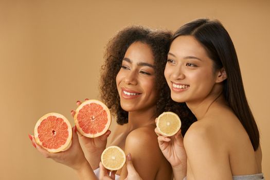 Portrait of two cute mixed race young women smiling aside, holding grapefruit and lemon cut in half while posing isolated over beige background. Health and beauty concept