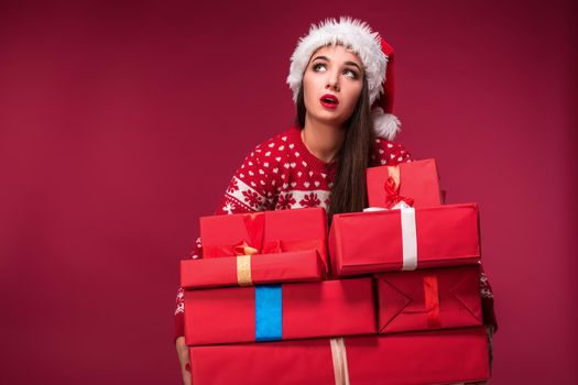 Young woman in sweater and christmas hat holding gift boxes on red background. Christmas sale. Emotional brunette. Copy space