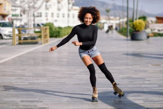Young fit black woman on roller skates riding outdoors on urban street. Smiling girl with afro hairstyle rollerblading on sunny day