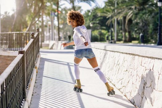 Young black woman on roller skates riding near the beach. Girl with afro hairstyle rollerblading on sunny promenade with sun backlight.