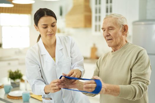 Caring nurse in a white coat explaining to a pensioner how to do exercises at home with fitness rubber