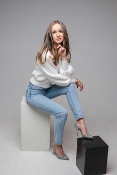 Studio full length portrait of pretty blonde woman in light blue jeans, fashionable heels and white pullover sitting on white cube and leaning on black with her leg.