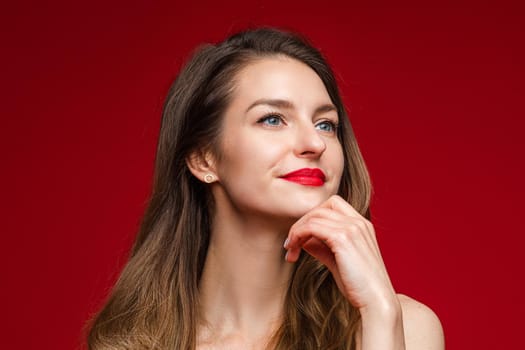 Portrait of gorgeous Caucasian woman with brown hair and red lips looking away pensively and holding hand at chin. Cutout on red background.