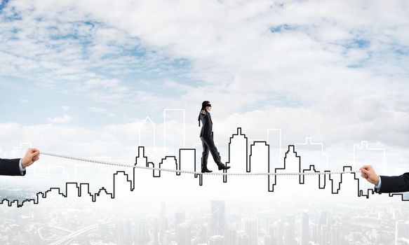 Businessman with blindfolder on eyes walking on rope over cityscape background