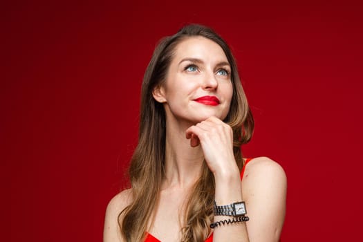 Portrait of gorgeous Caucasian woman with brown hair and red lips looking away pensively and holding hand at chin. Cutout on red background.