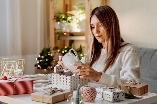 Woman wrapping Christmas eco natural gift boxes at home. Preparing presents on white table with decor elements, lights and items Christmas or New year DIY packing Concept.