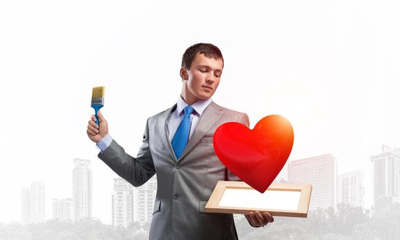 Creative businessman painter holding paint brush and 3d red heart. Portrait of young man in business suit and tie on cityscape background. Happy valentine day. Falling love concept.