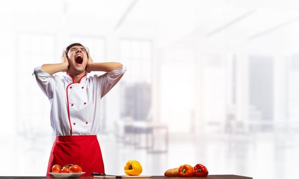 Young male chef screaming in shock and horror. Emotional caucasian chef in white hat and red apron standing with open mouth in light kitchen interior. Creative marketing campaign, big discounts.