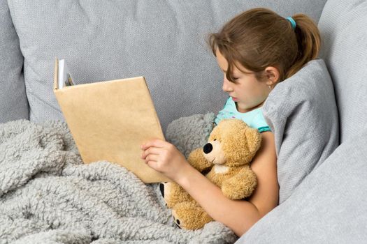 Beautiful girl sitting on comfortable sofa with teddy bear and reading book. Portrait of preteen girl with book and toy covered with plaid. Child resting on couch hugging favorite stuffed toy