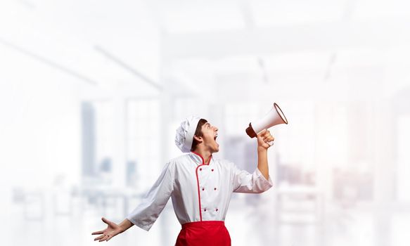 Young chef shouting loudly into megaphone. Emotional caucasian chef in white hat and red apron on light blurred background. Restaurant advertisement, food retail campaign announcement.
