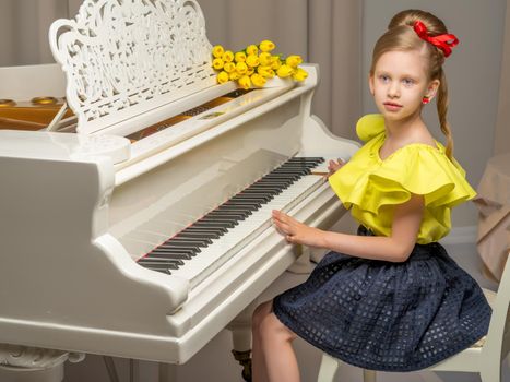 A sweet schoolgirl girl sits beside a white grand piano on which lies a bouquet of flowers. The concept of studying music, spring mood, happy people.