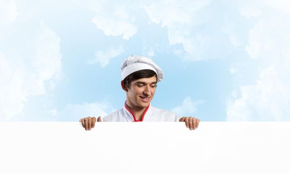 Young male chef with blank signboard. Handsome chef in white and red uniform on blue sky background. Cooking classes announcement. Professional catering service concept with copy space.