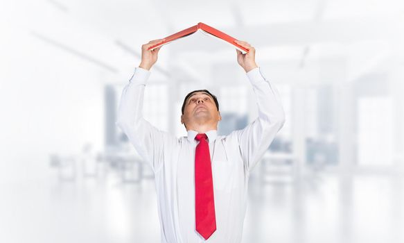Senior businessman holding open book overhead as roof. Portrait of adult man in white shirt and tie standing in blurred office interior. Education and professional knowledges. Reference information.