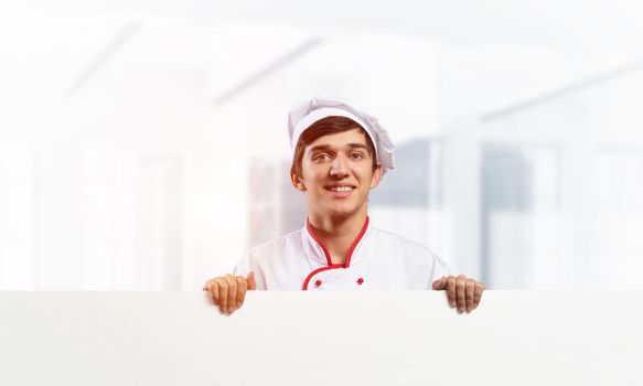 Young male chef with blank white signboard. Handsome chef in white and red uniform on light room blurred background. Cooking classes announcement. Professional catering service concept with copy space