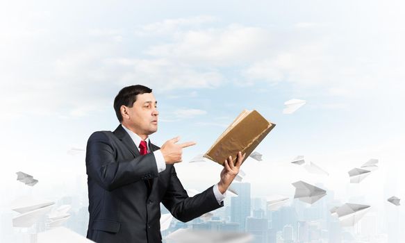 Senior businessman finger pointing into open book. Man in business suit and tie standing on cityscape background with flying paper airplanes. Manager holding handbook. Reference information concept