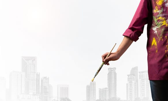Close up artist hand holding paintbrush. Painter in shirt standing on background modern skyscrapers. Creative hobby and artistic occupation concept with copy space. Art imagination and inspiration