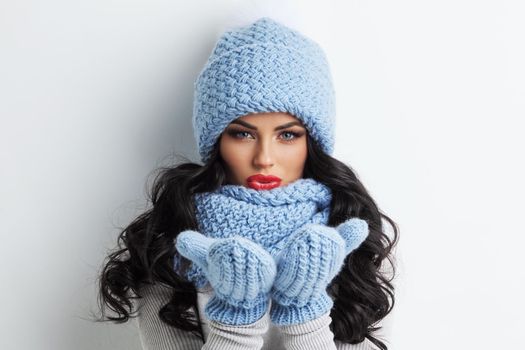 Beautiful woman in blue winter hat, scarf and mittens blowing on palms, white background